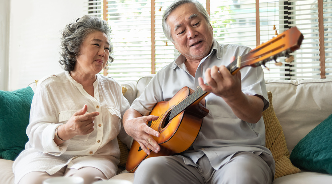 An older man plays guitar and woman is seated beside him