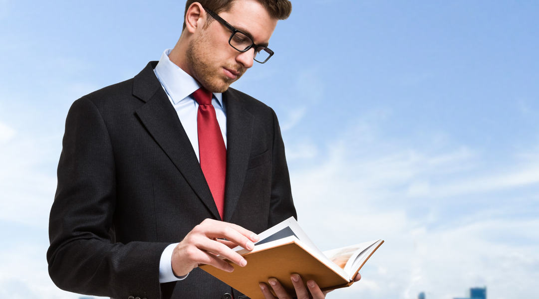 Man standing in black suit looks down at book he's holding