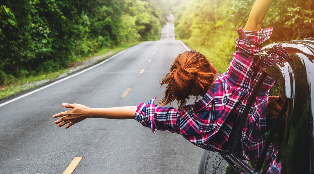 Woman hanging out of car along side of long road with trees