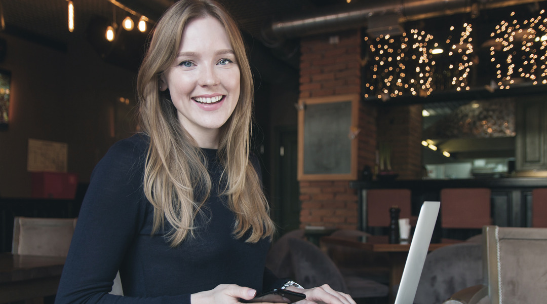 Smiling woman with cell phone and laptop in coffee shop