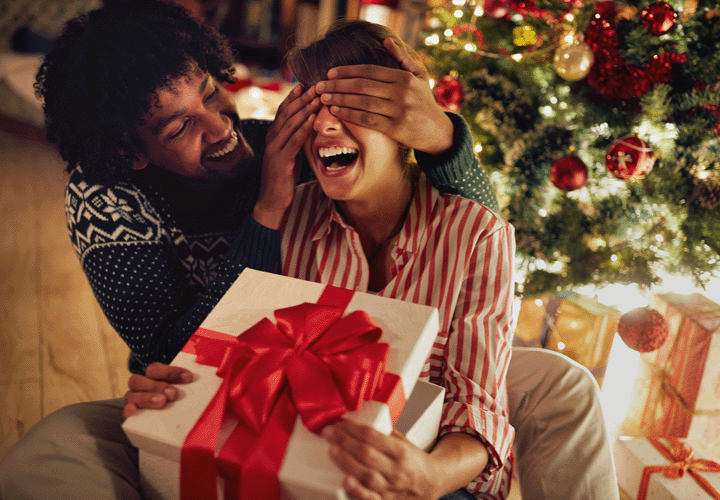 Man holds his hands over a woman's eyes to surprise her with a wrapped present she's holding