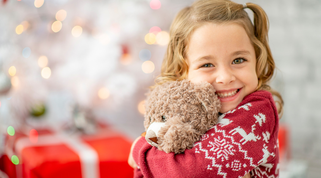 A smiling young girl holds a teddy bear in front of presents.