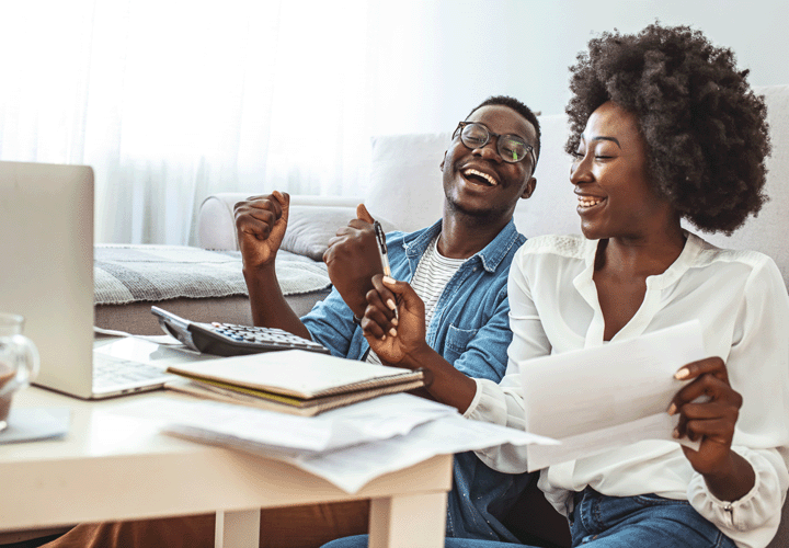 A man and a woman are seated on the floor using the coffee table as a desk. They're happy, sharing a laugh as their laptop and paperwork surround them.